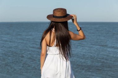 Beyaz elbise ve güneşli bir deniz kenarında kahverengi şapka stand uzun saçlı güzel esmer kız.