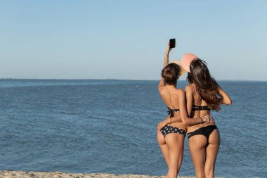 Arkadan görünüm mayolar iki ince esmer kız ve kapaklar kucaklama içinde güneşli bir günde denize yakın bir selfie yapmak.