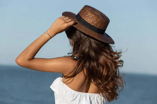 Retrato de linda menina de cabelos escuros em vestido branco e chapéu perto do mar em um dia ensolarado. Visão traseira . — Fotografia de Stock