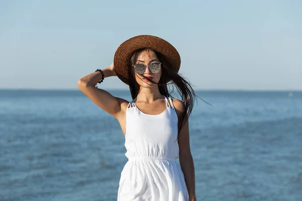 Menina deslumbrante de cabelos escuros em vestido branco, óculos de sol e chapéu marrom perto do mar em um dia ensolarado . — Fotografia de Stock