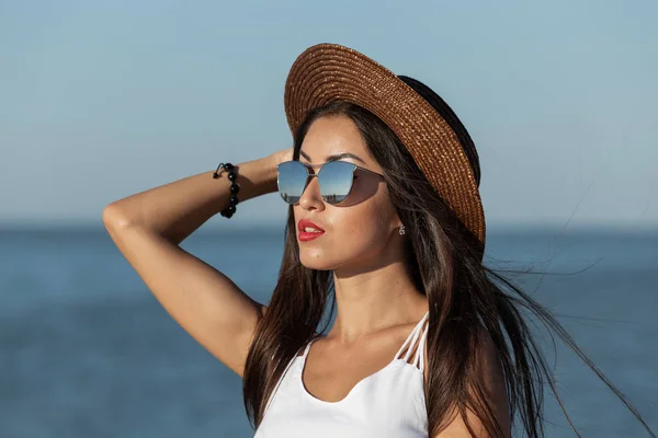 Retrato de uma linda garota morena com cabelos longos e batom vermelho em vestido branco, óculos de sol e chapéu marrom perto do mar — Fotografia de Stock