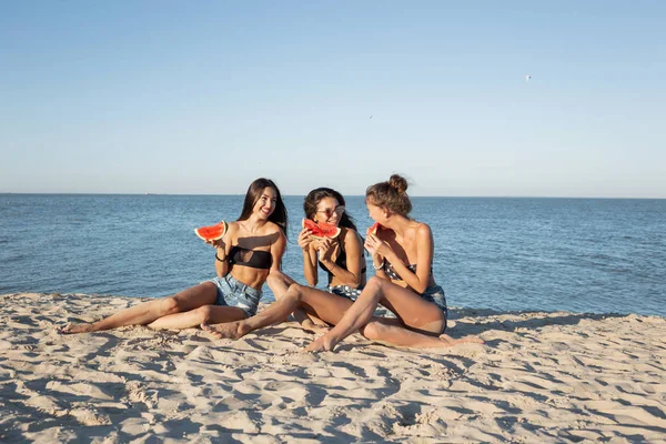 Três meninas bonitas em shorts jeans e sutiãs pretos estão sentados na areia perto do mar, segurando fatias de melancia e sorrindo . — Fotografia de Stock