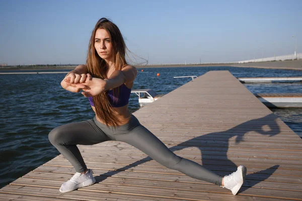 Hermosa joven con el pelo largo y castaño vestido con ropa deportiva haciendo estiramiento en el muelle de madera en el día de verano  . — Foto de Stock