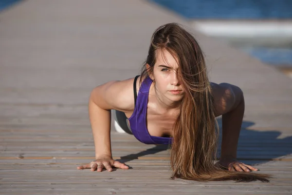 Hermosa joven con el pelo largo vestido con ropa deportiva está haciendo flexiones en el muelle de madera en el día de verano — Foto de Stock