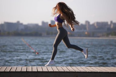 Uzun kahverengi saçlı bir spor üniforma giymiş güzel genç kız yaz gününde ahşap iskele üzerinde çalışıyor .