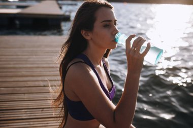 Spor top bir uzun kahverengi saçlı Genç güzel kız ahşap iskele üzerinde oturur ve suyu içer .