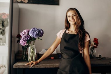 Bir vazo ile mavi ve Lila ortanca tabloyla yakınındaki bir siyah önlük çiçekçi kız duruyor