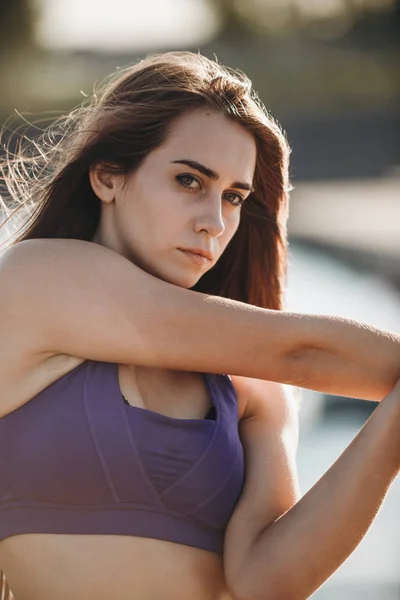 Giovane bella ragazza con lunghi capelli castani in un top sportivo sta facendo stretching delle braccia sul molo di legno  . — Foto Stock