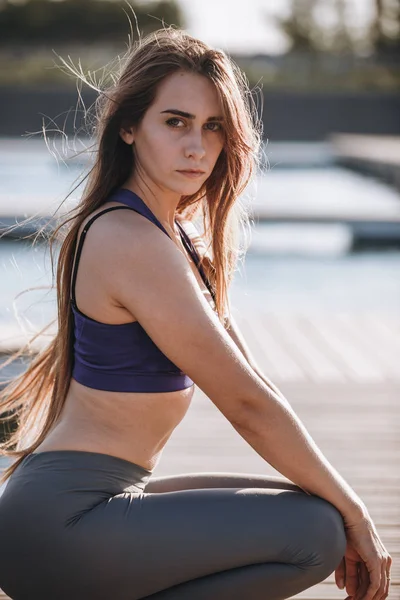 Joven hermosa chica con el pelo largo y castaño en un top deportivo y medias está en cuclillas en el muelle de madera  . — Foto de Stock