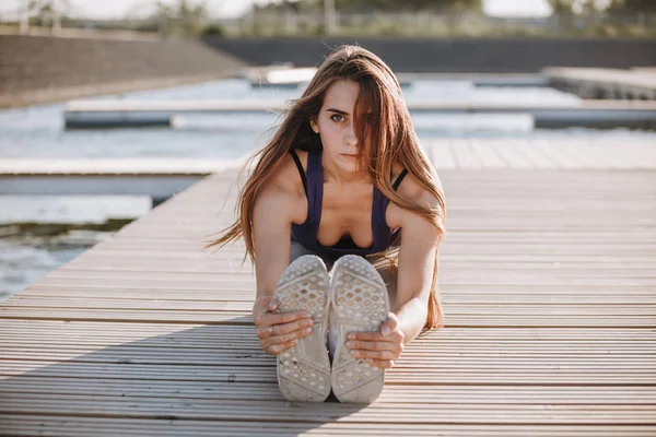 Joven chica delgada con el pelo castaño largo en un top deportivo y medias haciendo estiramiento en el muelle de madera  . — Foto de Stock