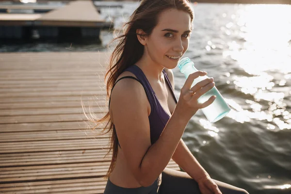 Mooi meisje met lang bruin haar in een sports shirt zit op de houten pier en water drinkt — Stockfoto