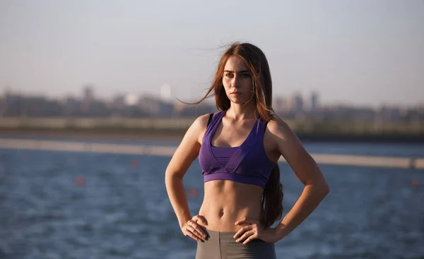 Mooi slank meisje met lang bruin haar in een sport-top staat in de buurt van de rivier . — Stockfoto