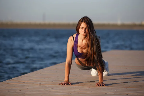 Mooi jong meisje met lang bruin haar gekleed in sport kleding plank op de houten pier op het water te doen op de zomerdag . — Stockfoto