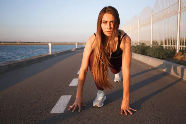 Joven hermosa chica con el pelo largo y castaño vestido con ropa deportiva haciendo estiramiento en la carretera a lo largo del embalse en el día soleado — Foto de Stock