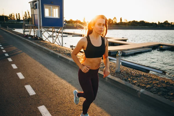 Hermosa joven con el pelo castaño largo vestido con ropa deportiva corre en el camino a lo largo del agua en la puesta del sol — Foto de Stock
