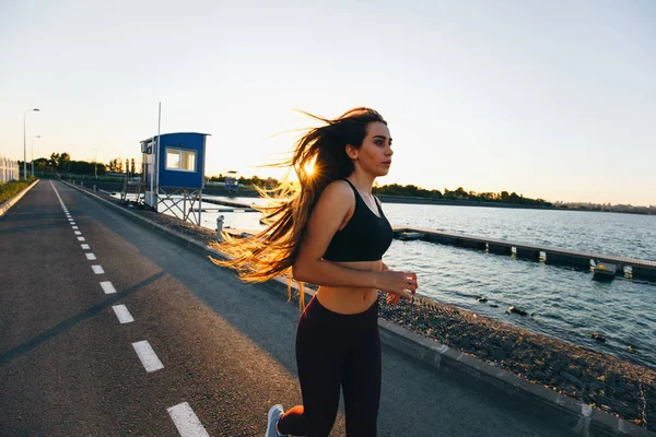 Hermosa joven con el pelo castaño largo vestido con ropa deportiva corre en el camino a lo largo del agua en la puesta del sol — Foto de Stock