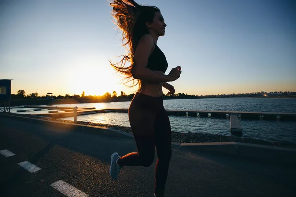 Magnifique jeune fille aux longs cheveux bruns vêtus de vêtements de sport court sur la route le long du réservoir au coucher du soleil — Photo