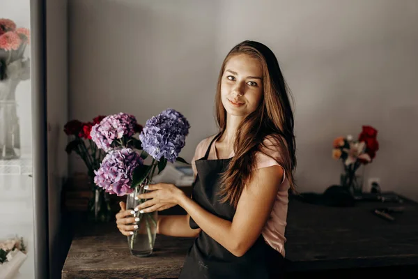 Дівчина-флорист в чорному фартусі тримає вазу з синьою і бузковою гортензією — стокове фото