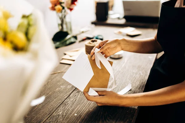 Florista está embalando um cartão postal em um envelope com fita branca na mesa de madeira — Fotografia de Stock