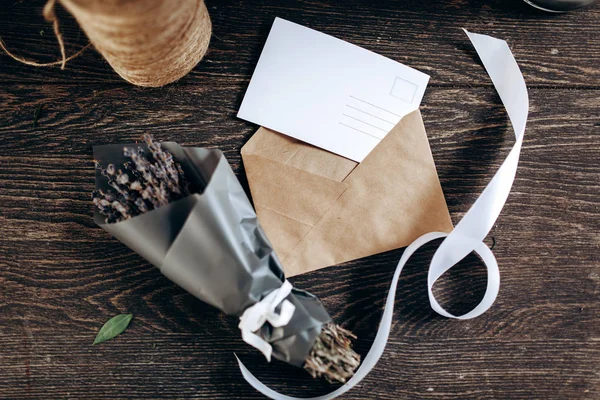 Un petit bouquet de fleurs emballées dans du papier foncé, carte postale, enveloppe et ruban blanc sont sur la table en bois — Photo