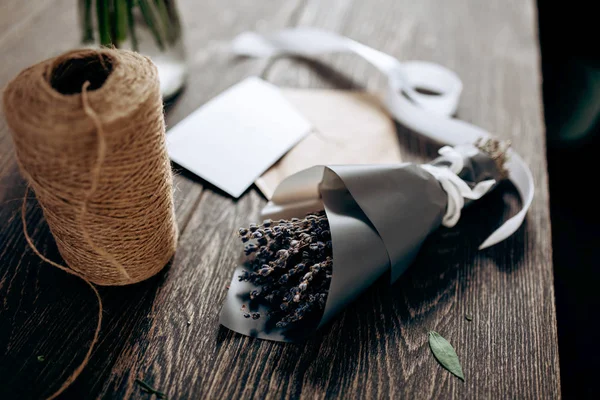 Un petit bouquet de fleurs emballé dans du papier foncé, carte postale, enveloppe, bobine de ficelle et ruban blanc sont sur la table en bois — Photo