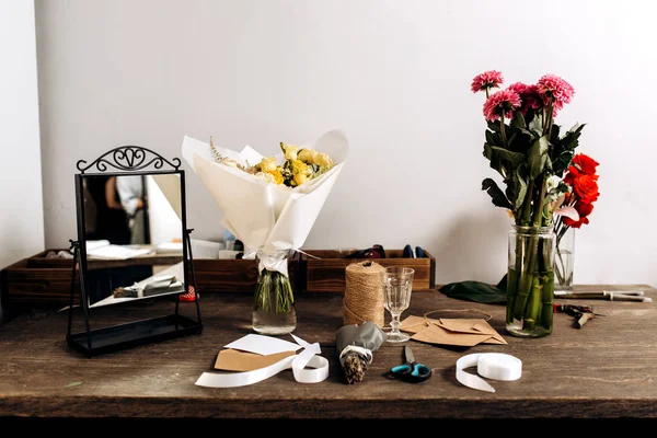 Il y a trois bouquets différents, miroir, carte postale, enveloppes, bobine de ficelle, ciseaux et ruban blanc sur la table en bois — Photo