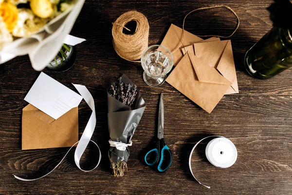 Un petit bouquet de fleurs emballé dans du papier foncé, carte postale, enveloppes, bobine de ficelle, ciseaux et ruban blanc sont sur la table en bois — Photo