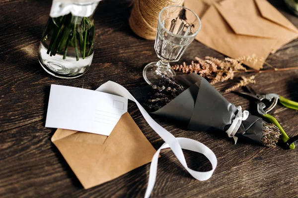 Il y a un petit bouquet de fleurs, verre à vin, carte postale, enveloppes et ruban blanc sur la table en bois — Photo