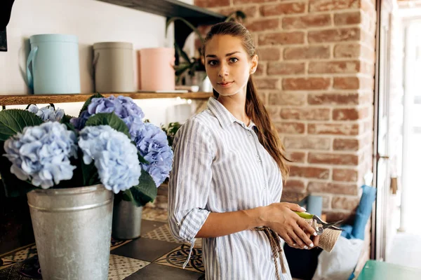 Menina morena bonita vestida com um vestido listrado fica na loja de flores perto do vaso com hortensia azul claro — Fotografia de Stock