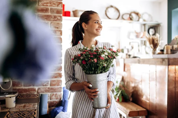 Красива брюнетка, одягнена в смугасту сукню, тримає вазу з рожевими і білими хризантемами в квітковому магазині — стокове фото