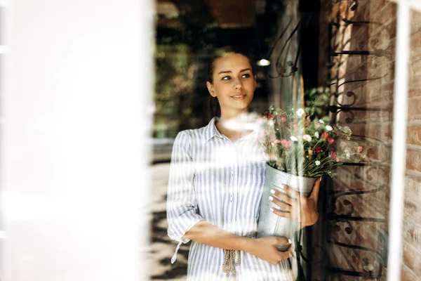 Menina morena bonita vestida com um vestido listrado segura um vaso com crisântemos rosa e branco à luz do sol — Fotografia de Stock