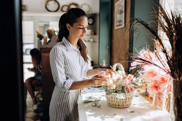 Bloemist gekleed in een gestreepte jurk collecties een boeket in de mand van verse bloemen van pastel kleuren in de bloemenwinkel — Stockfoto