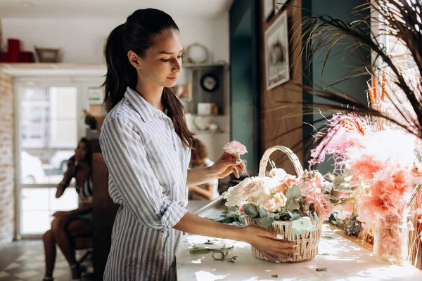 Bloemist gekleed in een gestreepte jurk collecties een boeket in de mand van verse bloemen van pastel kleuren in de bloemenwinkel — Stockfoto