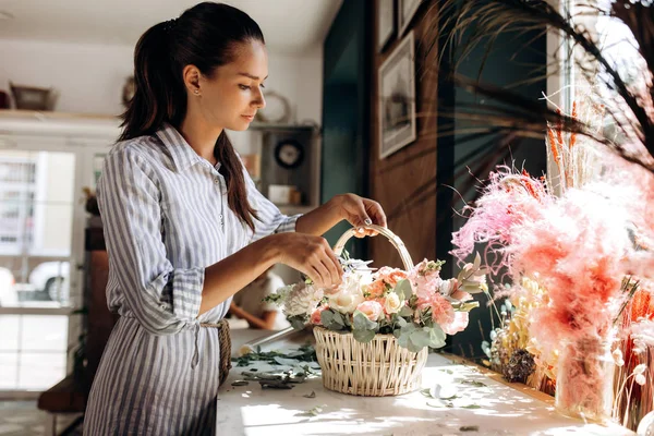 Флорист одягнений в смугасту сукню колекції букет в кошику зі свіжих квітів пастельних кольорів в квітковому магазині — стокове фото