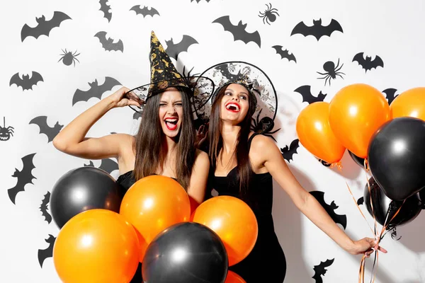 Dos encantadoras mujeres jóvenes con sombreros de brujas están con globos negros y naranjas sobre un fondo blanco con murciélagos negros. Halloween —  Fotos de Stock