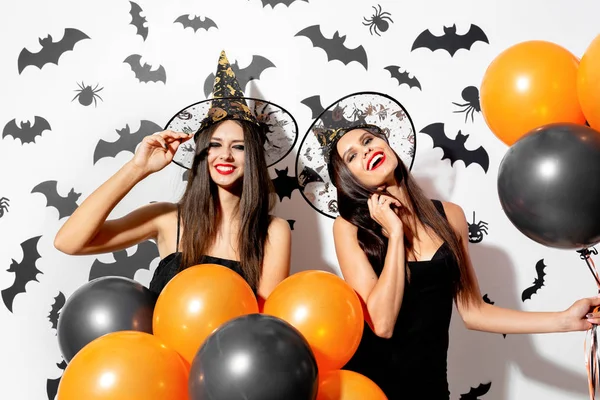 Due affascinanti giovani donne in cappelli da strega sono con palloncini neri e arancioni su uno sfondo bianco con pipistrelli neri. Halloween — Foto Stock