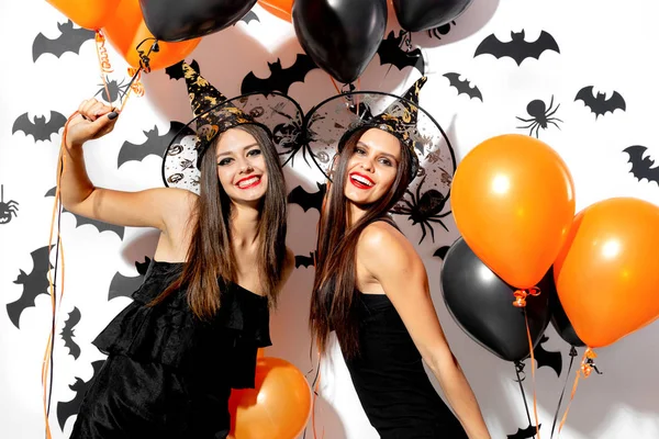 Due belle giovani donne in cappelli da strega sono con palloncini neri e arancioni su uno sfondo bianco con pipistrelli neri. Halloween — Foto Stock