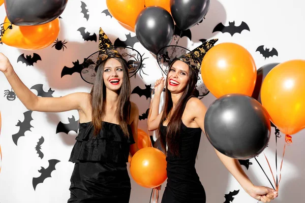 Zwei schöne junge Frauen mit Hexenhüten sind mit schwarzen und orangefarbenen Luftballons auf weißem Hintergrund mit schwarzen Fledermäusen. halloween — Stockfoto