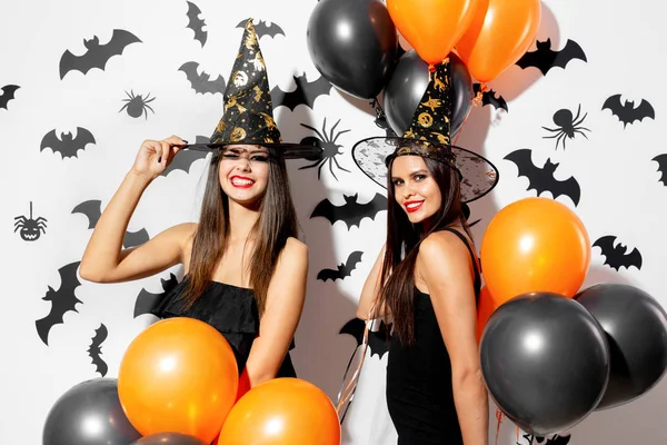 Zwei schöne junge Frauen mit Hexenhüten sind mit schwarzen und orangefarbenen Luftballons auf weißem Hintergrund mit schwarzen Fledermäusen. halloween — Stockfoto