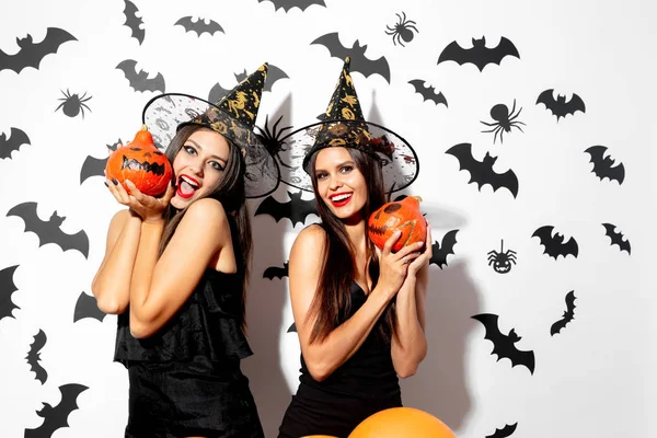 Zwei charmante brünette Frauen in schwarzen Kleidern und Hexenhüten halten Halloween-Kürbisse auf weißem Hintergrund mit Fledermäusen und Spinnen — Stockfoto