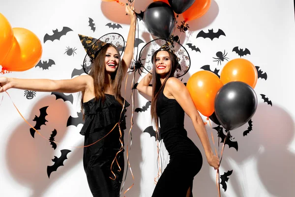 Två vackra brunett kvinnor i häxor hattar är med svart och orange ballonger på en vit bakgrund med fladdermöss och spindlar. — Stockfoto