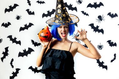 Cadılar Bayramı Balkabağı siyah yarasalar ve örümcekler beyaz bir arka plan üzerinde mavi bir peruk ve witchs şapkalı komik genç kadın tutar