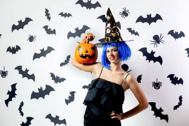 Cadılar Bayramı Balkabağı siyah yarasalar ve örümcekler beyaz bir arka plan üzerinde mavi bir peruk ve witchs şapkalı komik genç kadın tutar