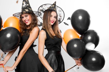 İki güzel kız cadı şapka ve siyah elbiseler siyah ve turuncu balonlarla iyi eğlenceler. Cadılar Bayramı
