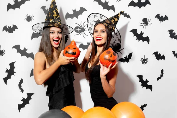 Zwei charmante brünette Frauen in schwarzen Kleidern und Hexenhüten halten Halloween-Kürbisse auf weißem Hintergrund mit Fledermäusen und Spinnen — Stockfoto