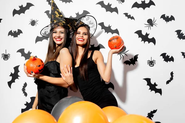 Dos hermosas mujeres morenas con sombreros de brujas se divierten con calabazas de Halloween sobre un fondo blanco con murciélagos y arañas —  Fotos de Stock