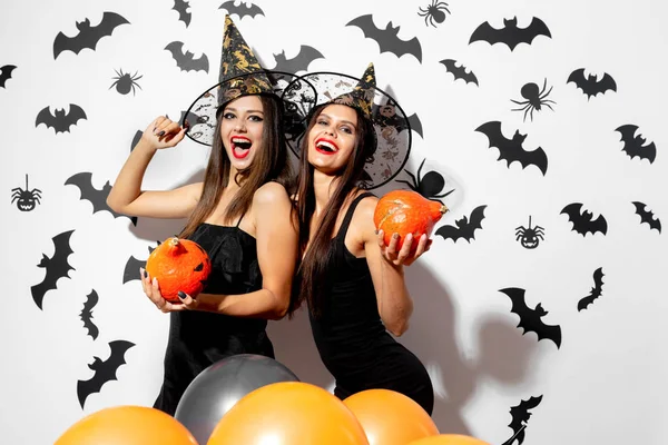 Due belle donne brune con cappelli da strega si divertono con le zucche di Halloween su uno sfondo bianco con pipistrelli e ragni — Foto Stock