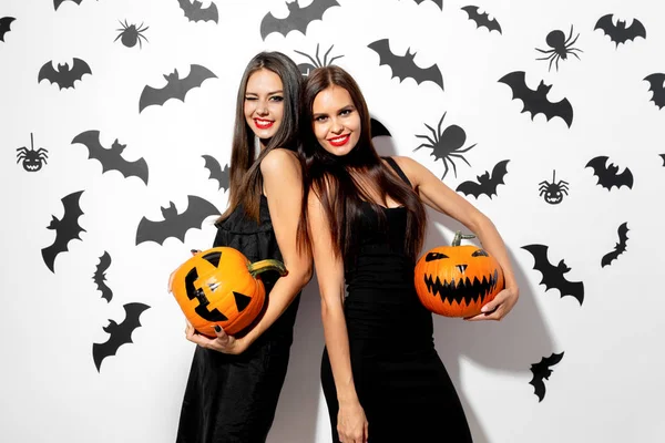 Zwei schöne brünette Frauen in schwarzen Kleidern haben Spaß mit Jack-o-Laternen auf weißem Hintergrund mit Fledermäusen und Spinnen — Stockfoto