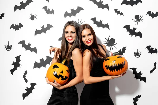 Zwei schöne brünette Frauen in schwarzen Kleidern haben Spaß mit Jack-o-Laternen auf weißem Hintergrund mit Fledermäusen und Spinnen — Stockfoto