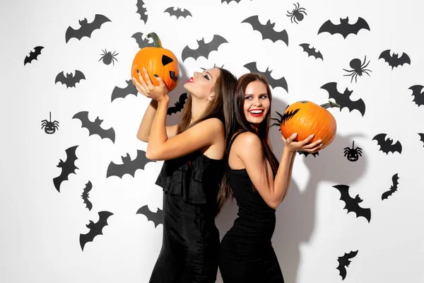 Zwei charmante brünette Frauen in schwarzen Kleidern mit rotem Lippenstift halten Halloween-Kürbisse auf weißem Hintergrund mit Fledermäusen und Spinnen — Stockfoto